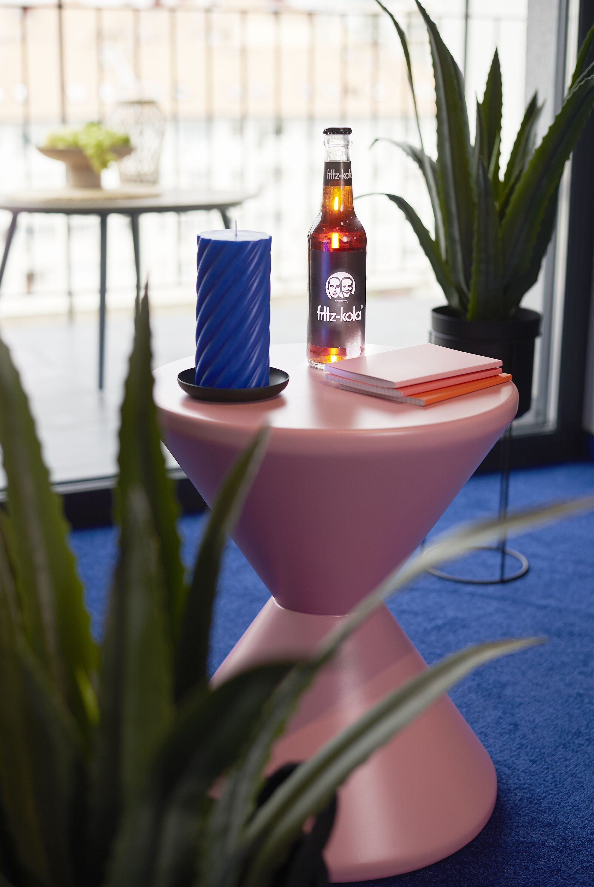 A pink table with a bottle of Fritz-Kola, a blue candle, and a notebook on it.