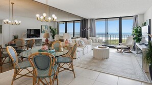 A spacious living room with a large window overlooking the beach, a dining area with wicker chairs, and a modern chandelier. - Beachfront 2BR/2BA South Seas condo with pool, WiFi, and beach access (Marco Island)
