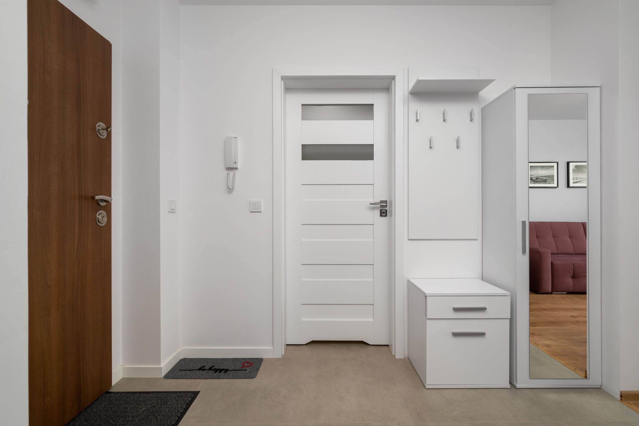 Un couloir avec une porte en bois, une porte blanche à lamelles horizontales, un meuble blanc avec des tiroirs et un miroir qui reflète une pièce où se trouve un canapé rose.