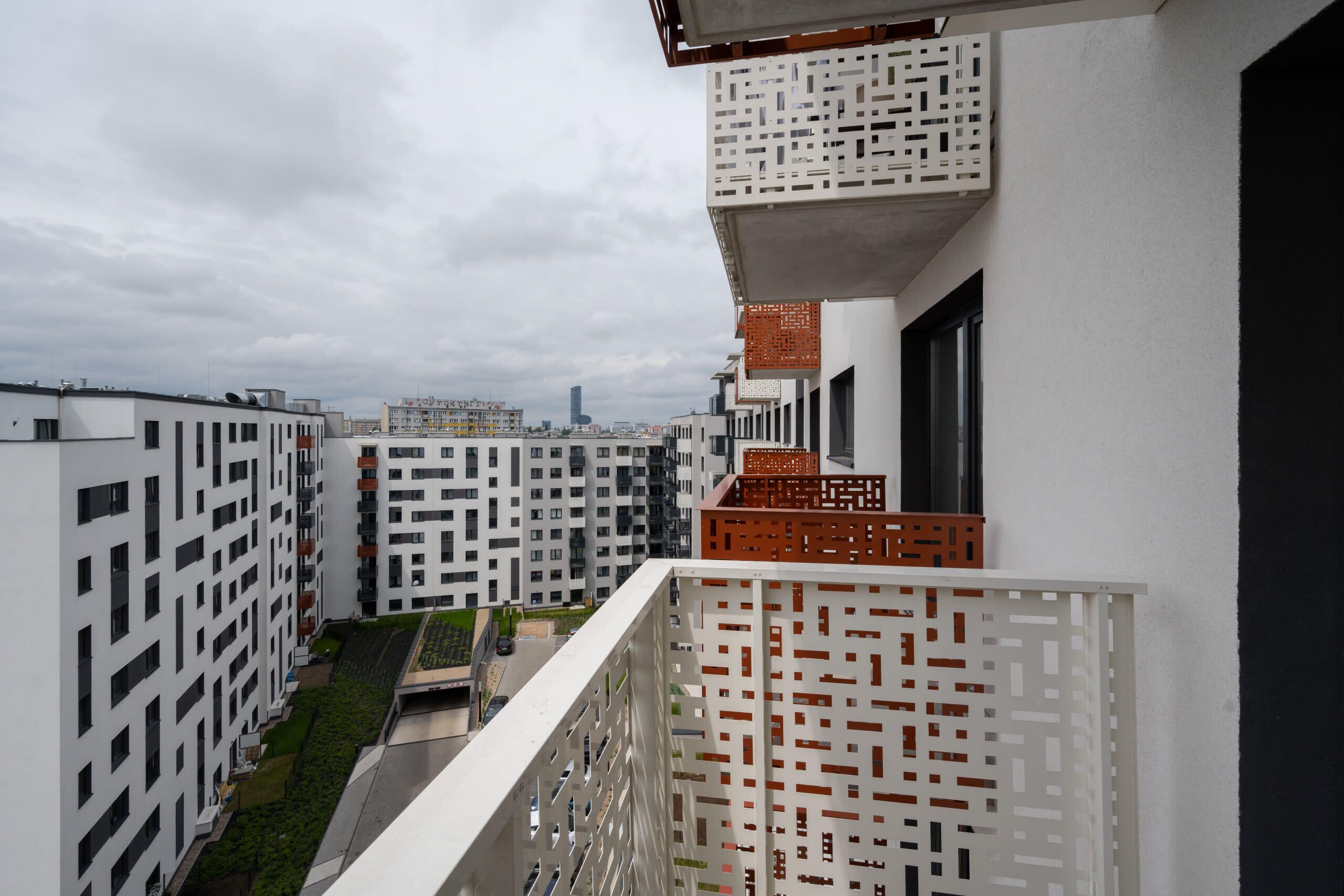 Un moderno edificio de apartamentos con fachada geométrica, balcones con barandillas ornamentadas y vistas de la ciudad al fondo.