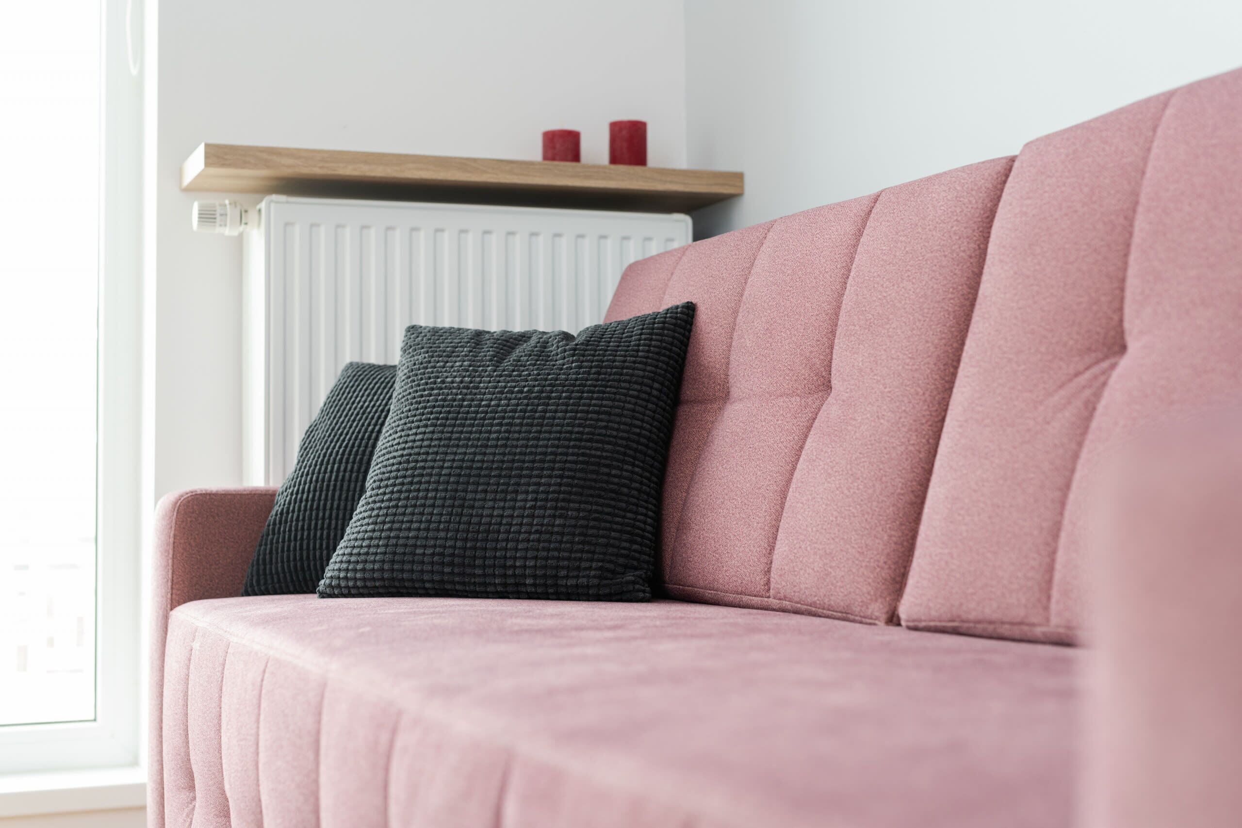 Een roze bank met twee donkere kussens, een houten plank met twee rode kaarsen, en een witte radiator op de achtergrond.