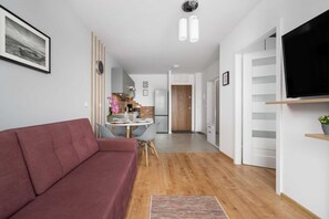 Ein modernes Wohnzimmer mit einer Couch, einem Essbereich mit Stühlen, einer Küche im Hintergrund und einem an der Wand montierten Fernseher.