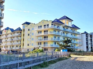 A multi-story hotel with balconies, a sandy beach in the foreground, and a clear sky. - Popular Adagio building, mid-town on the ocean! (Ocean City)