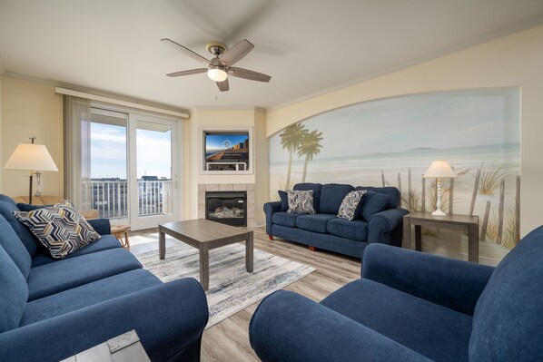 A living room with a blue sofa set, a fireplace, a ceiling fan, and a beach mural on the wall. - Popular Adagio building, mid-town on the ocean! (Ocean City)