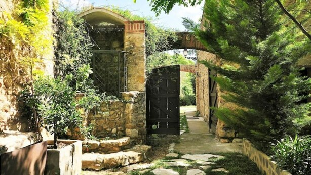 Un sendero de piedra que atraviesa un jardín rústico con una verja negra, rodeado de follaje y muros de piedra.