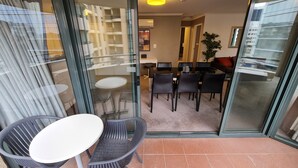 A modern living room with a glass door leading to a balcony. Inside, there is a dining area with a table and chairs, a sofa, and a potted plant.