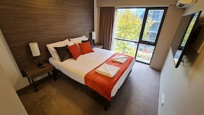 A hotel room with a large bed, a nightstand, a lamp, a TV, and a window with a view of trees.