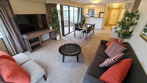 A modern living room with a flat-screen TV, a sofa set, a dining area, and a kitchen in the background.