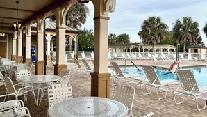 An outdoor pool area with white lounge chairs and tables, surrounded by palm trees and covered by a pergola with columns. - Sun Splashed - 3/2 w/ Golf Cart in The Village of Amelia (The Villages)
