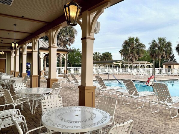 An outdoor pool area with white lounge chairs and tables, surrounded by palm trees and covered by a pergola with columns.