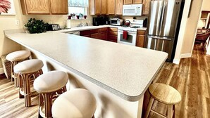 A modern kitchen with wooden cabinets, a large island with bar stools, stainless steel appliances, and a window with lace curtains. - Sun Splashed - 3/2 w/ Golf Cart in The Village of Amelia (The Villages)