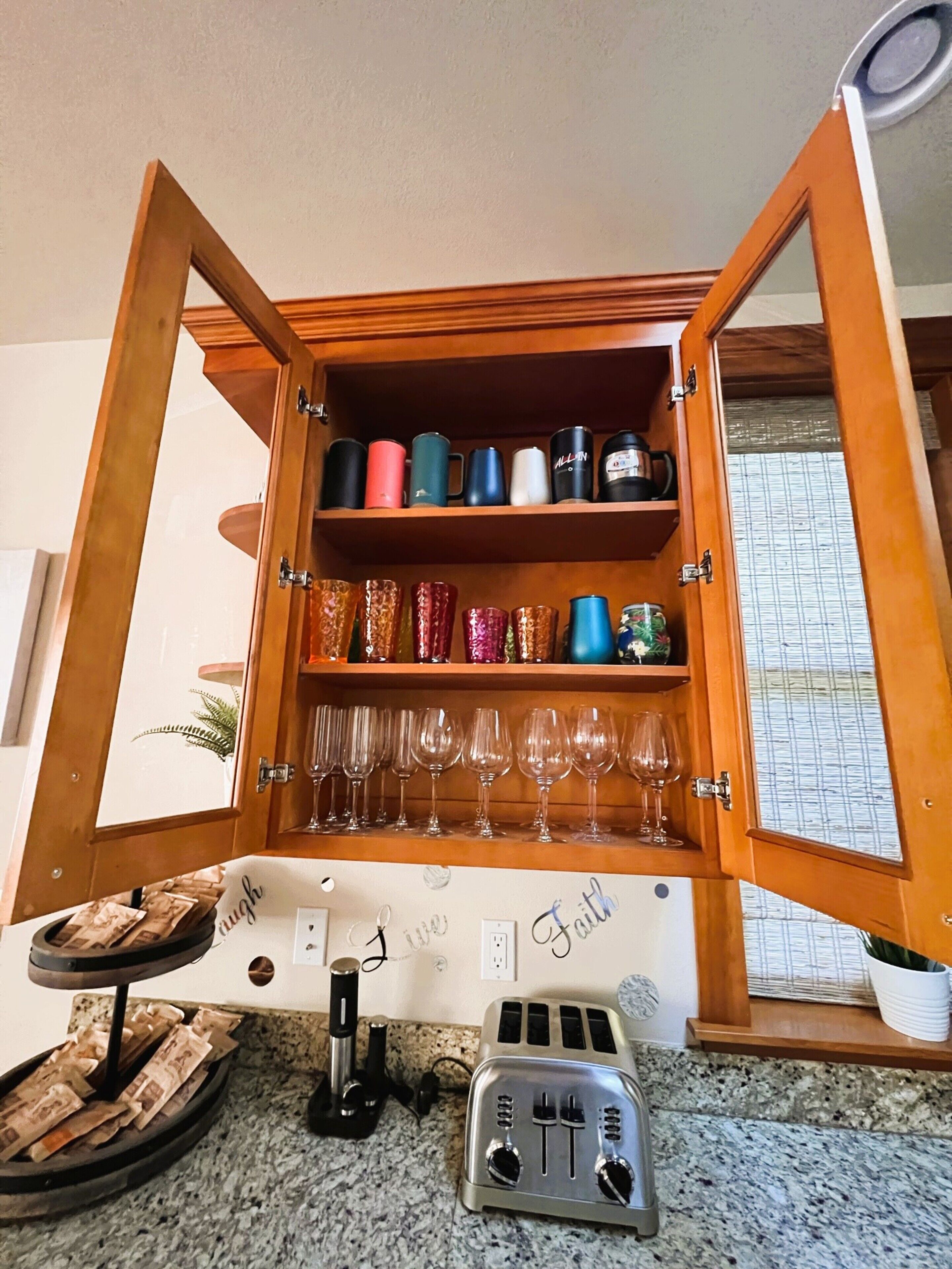 A kitchen with a wooden cabinet containing colorful mugs and wine glasses, a toaster, and a coffee maker on the counter.