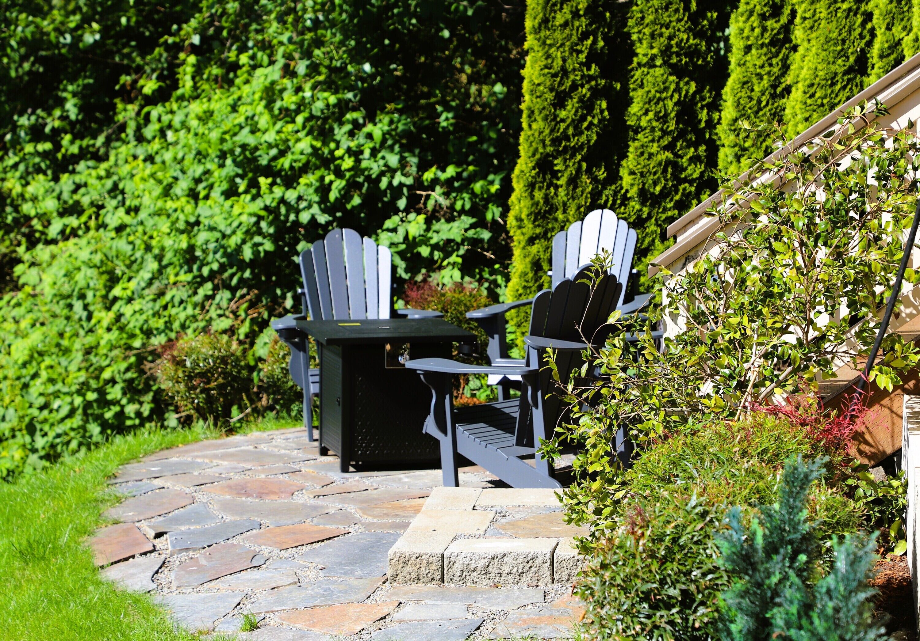 Une terrasse aménagée dans un jardin, avec des fauteuils Adirondack, un chemin de pierres et une végétation luxuriante.