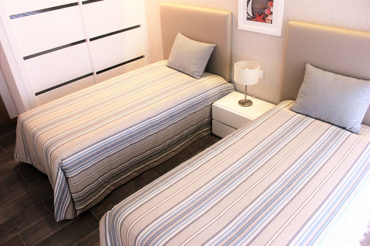 A hotel room with two beds, each with striped bedding, a bedside table with a lamp, and a wall-mounted storage unit.