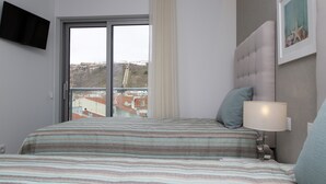 A hotel room with a bed, a television mounted on the wall, a window with a view of buildings, and a framed picture on the wall. - Apartamentos Turísticos da Nazaré (Nazaré)