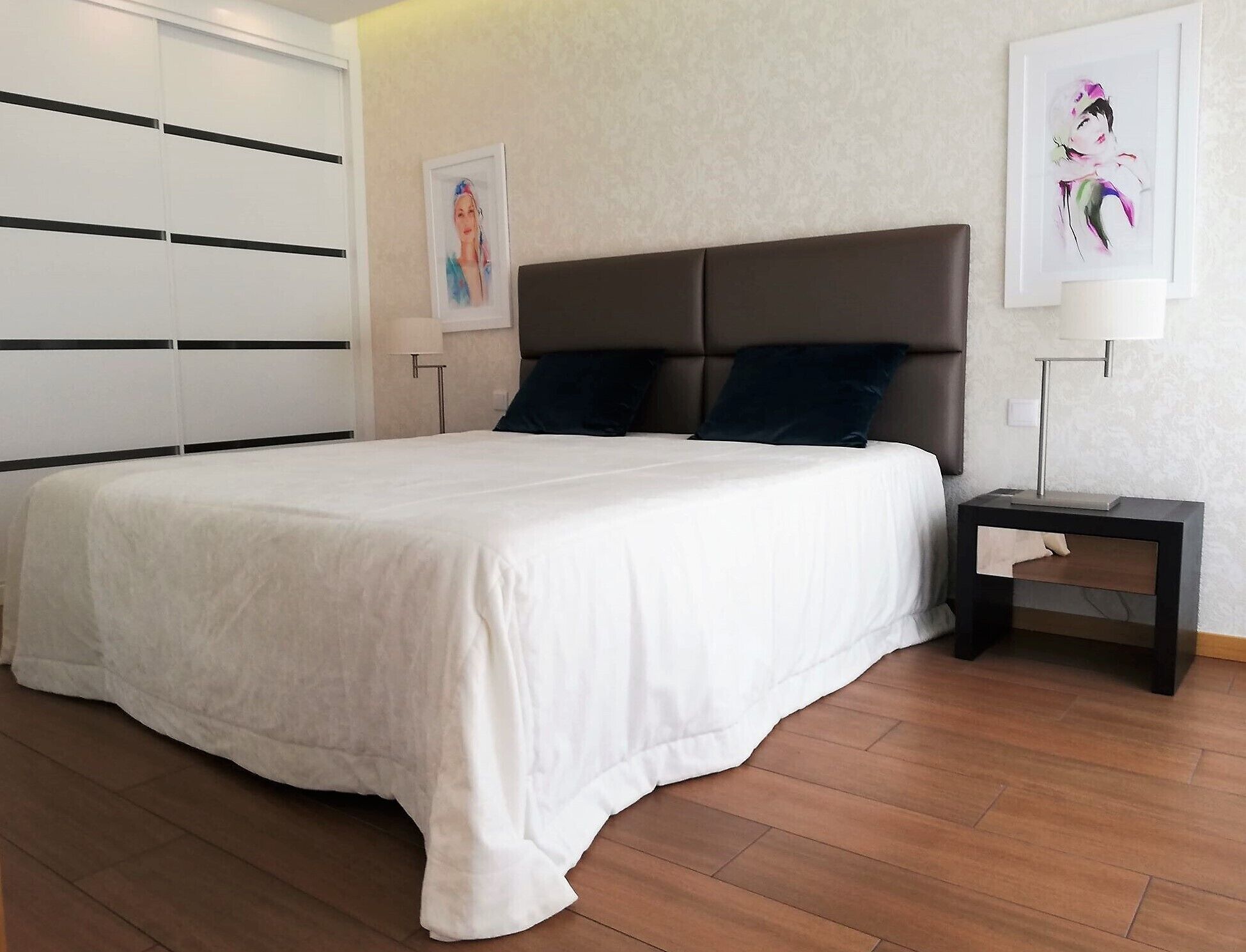 Un dormitorio con una cama grande, un cabecero oscuro, dos almohadas, una mesita de noche con una lámpara y una obra de arte enmarcada en la pared.