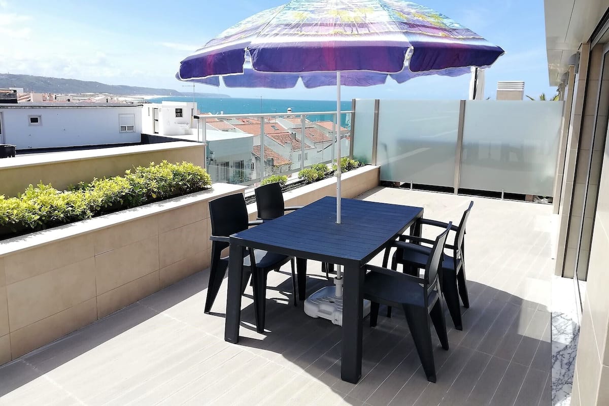 A rooftop terrace with a table and chairs, a colorful umbrella, and a view of the city and sea.
