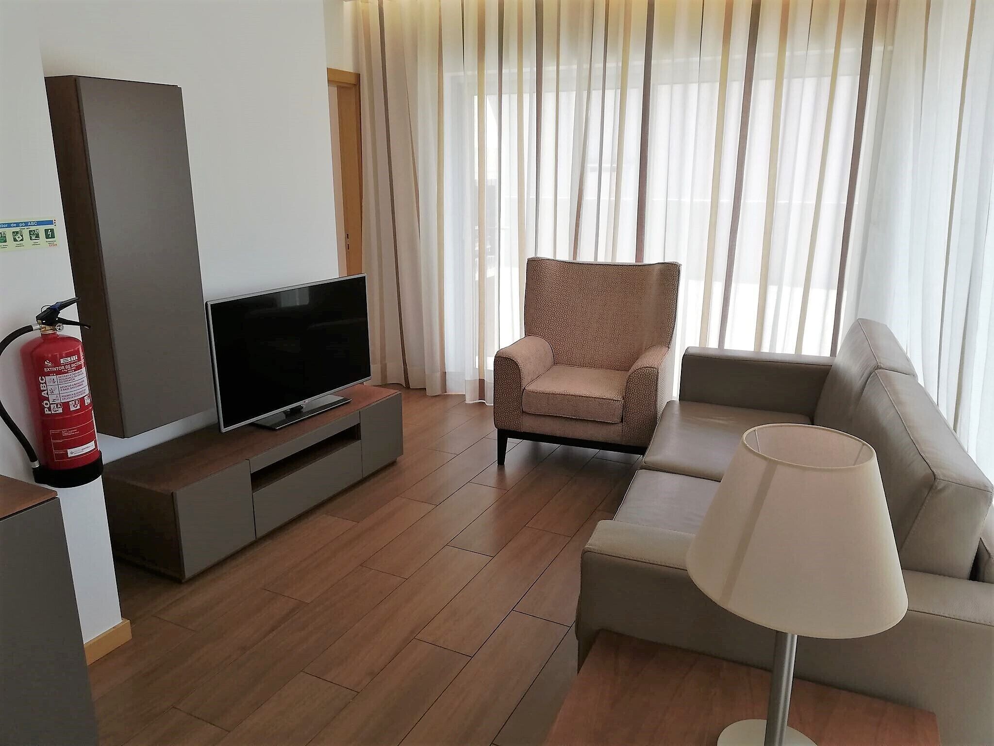 A modern living room with a flat-screen TV on a grey console, a beige armchair, a sofa, and a floor lamp.