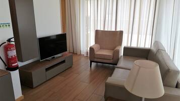 A modern living room with a flat-screen TV on a grey console, a beige armchair, a sofa, and a floor lamp.