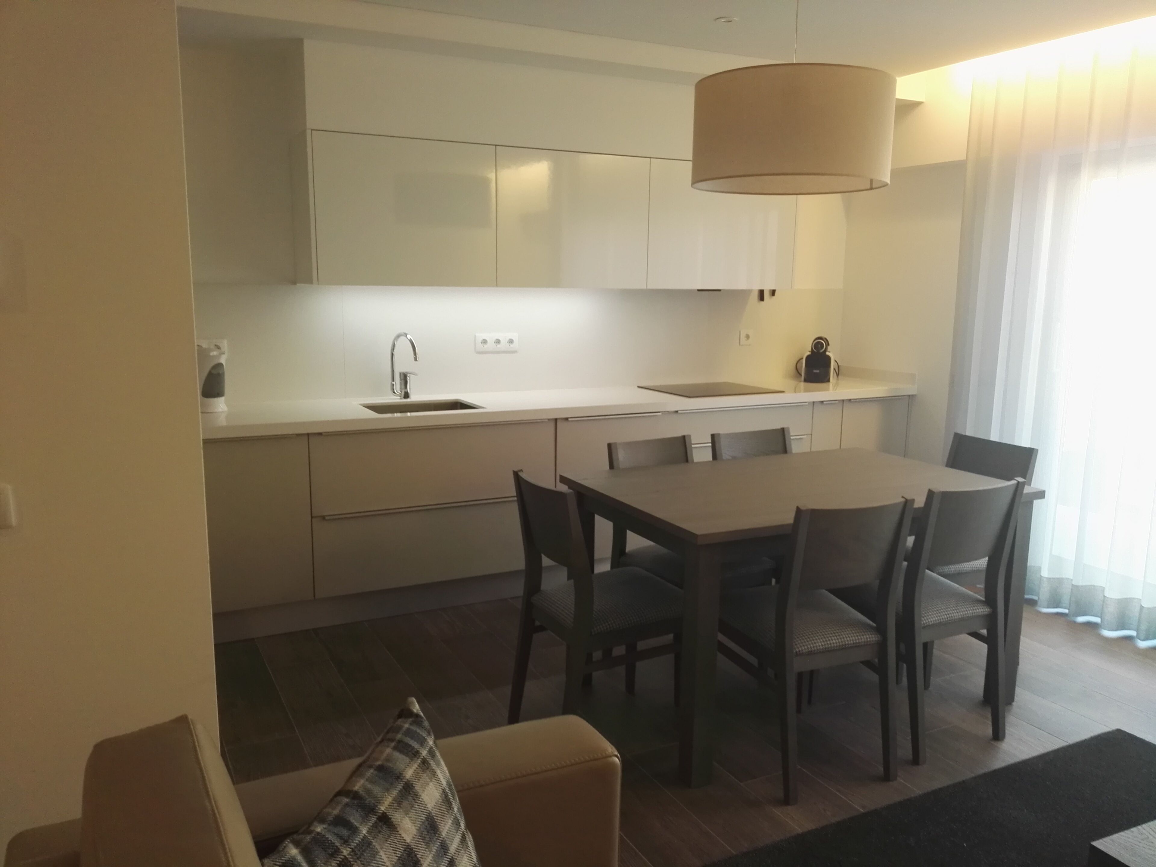 A modern kitchen with white cabinets, a dining table with chairs, a sofa with a plaid pillow, and a large window with sheer curtains.
