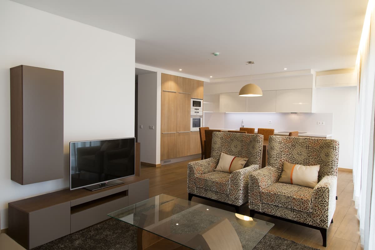 A modern living room with a flat-screen TV, two patterned armchairs, a glass coffee table, and a kitchen in the background.