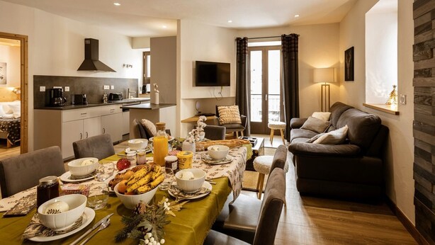 A modern living room with a sofa, a dining table set for breakfast, and a kitchen area with a stove and sink. - 4-room apartment, South facing, 6 people, pets allowed (Valloire)