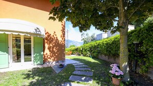 A garden with a stone pathway, a tree with flowers, and a building with green doors. - MALCESINE DREAM APARTMENT (Malcesine)