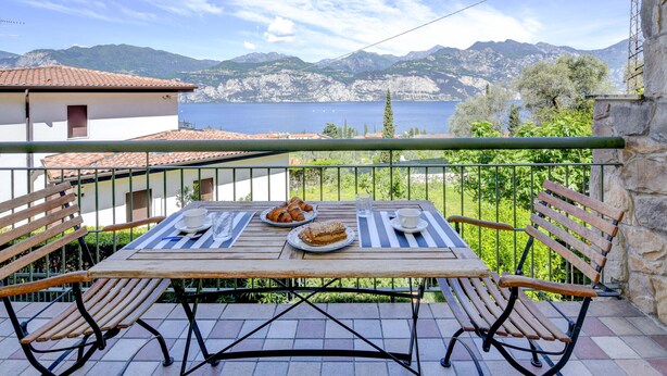 A balcony with a table set for two, overlooking a lake and mountains. - MALCESINE DREAM APARTMENT (Malcesine)