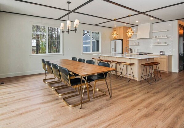 A spacious dining area with a long wooden table and chairs, a kitchen with white cabinets and a central island, and large windows offering a view of the outdoors.