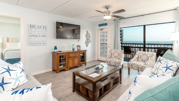 A living room with a view of the ocean, a ceiling fan, and a wooden cabinet. - Saida III 1105 (3115) - Coastal 2 bedroom Condo for 6 guests in Beachfront Resort with 3 pools and amazing View (South Padre Island)