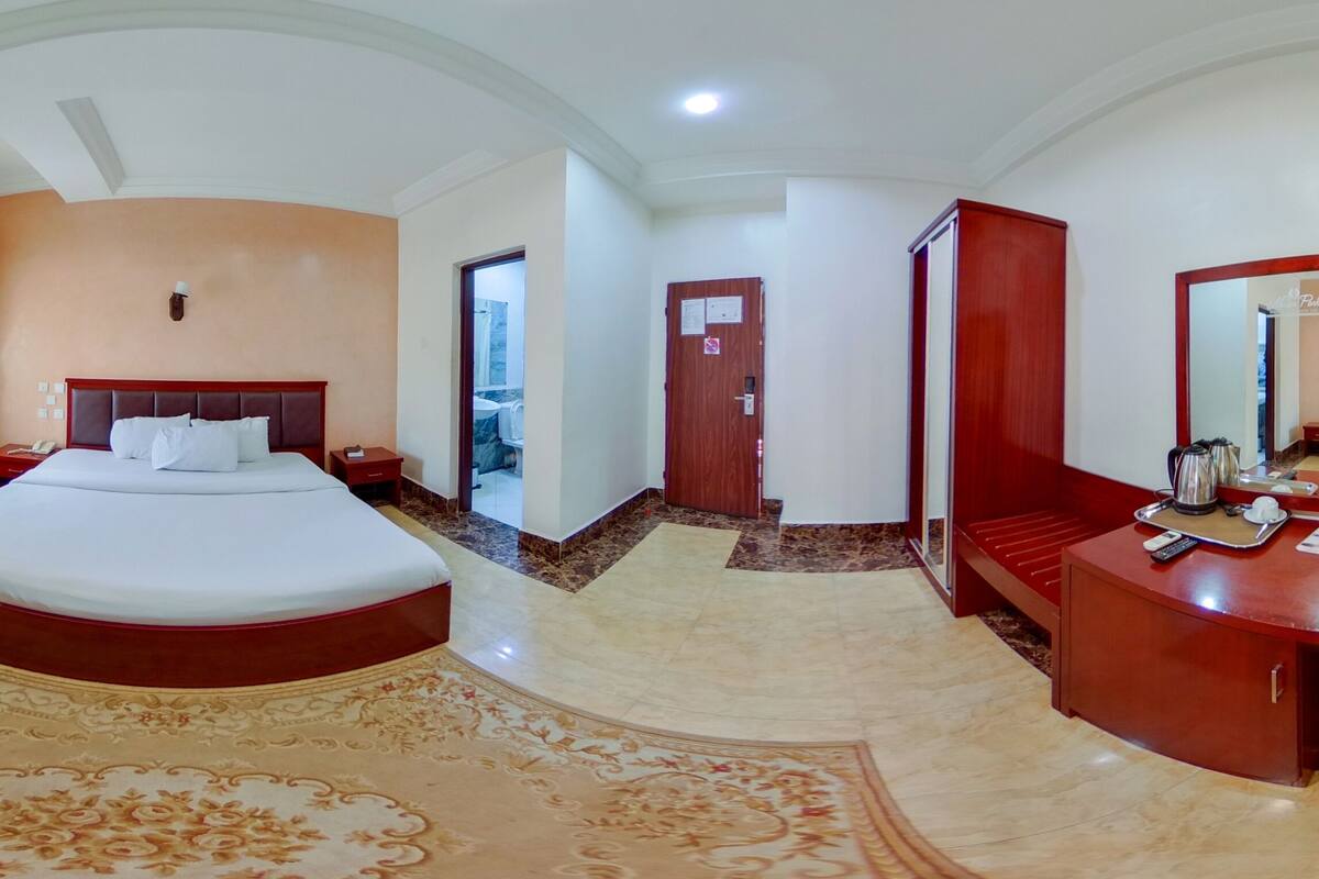 A hotel room with a large bed, a small table, a chair, a TV, and a bathroom visible through an archway.