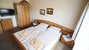 Una cama bien hecha con sábanas blancas y dos almohadas, una mesita de noche de madera con una lámpara, un armario de madera y un televisor en la pared.