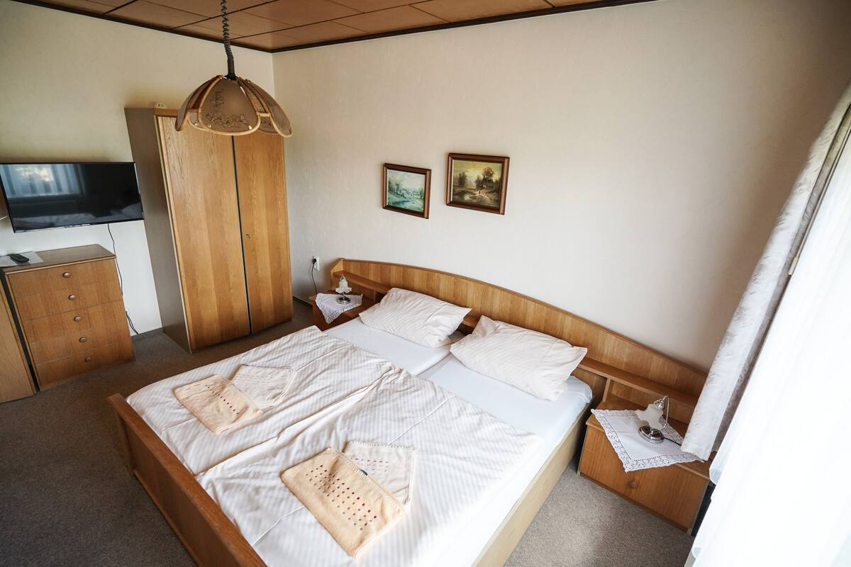 A neatly made bed with white linens and two pillows, a wooden nightstand with a lamp, a wooden wardrobe, and a television on the wall.