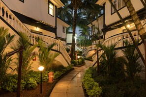 A well-lit, narrow pathway flanked by lush greenery and palm trees, leading to a building with a classic architectural style. - 2 Bedroom/2 Bath apartment in Candolim Beach resort (Candolim)