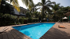 A swimming pool with wooden deck, surrounded by palm trees and a building.