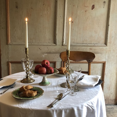 A table set with a white tablecloth, two lit candles, a plate of apples, a plate of croissants, and a green plate with a pastry.