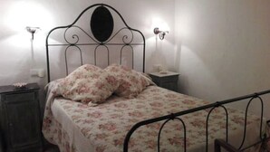 A single bed with a floral bedspread and two pillows, a bedside table with a lamp, and a wall-mounted light fixture. - Historical country house Costa Brava | Dali 2 Pax (Vilanant)