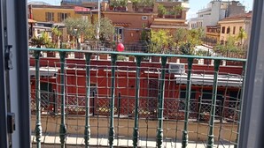 Vista a través de una ventana con reja metálica, mostrando edificios con balcones y antenas parabólicas, bajo un cielo azul despejado.