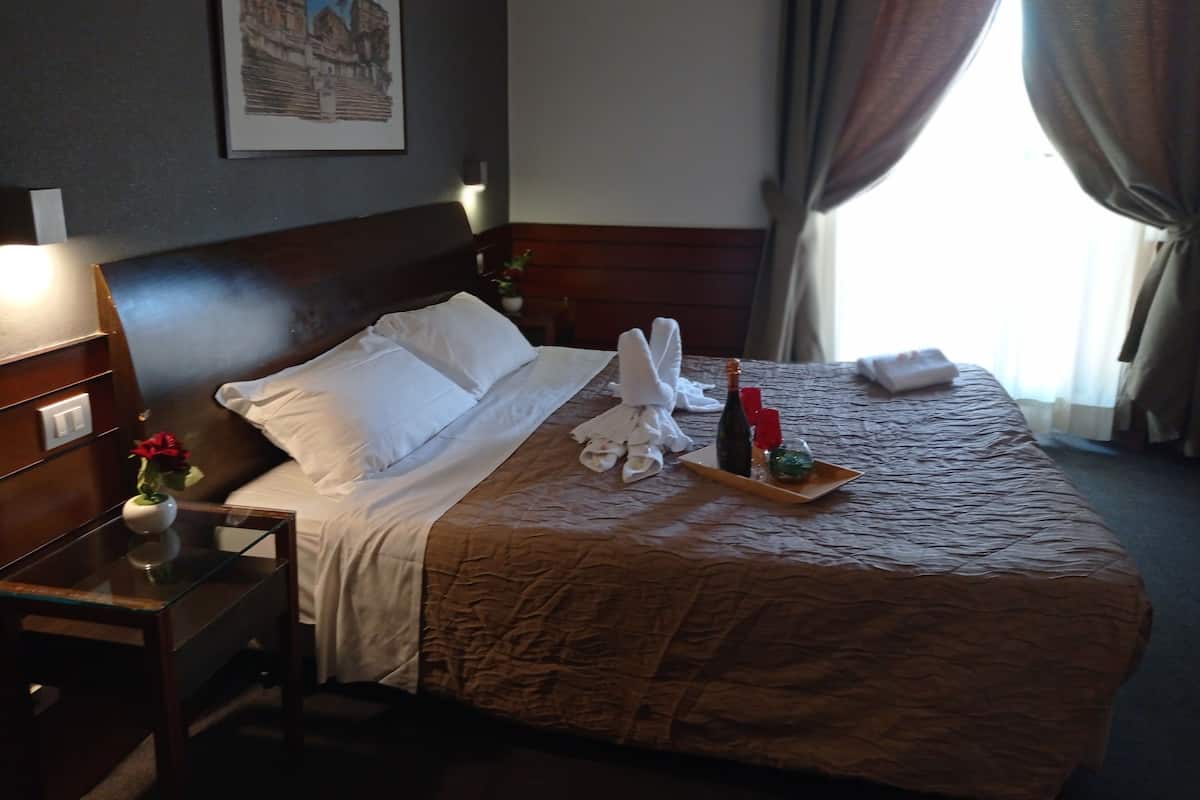 Una habitación de hotel con una cama bien hecha, una mesita de noche con un florero, un cuadro en la pared y una ventana con cortinas.
