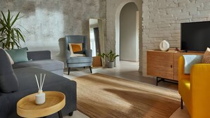 A modern living room with a grey sofa, a blue armchair, a yellow armchair, a wooden side table with a white vase, a TV on a wooden stand, and a white brick wall. - Butik Art Hotel (Budapest)