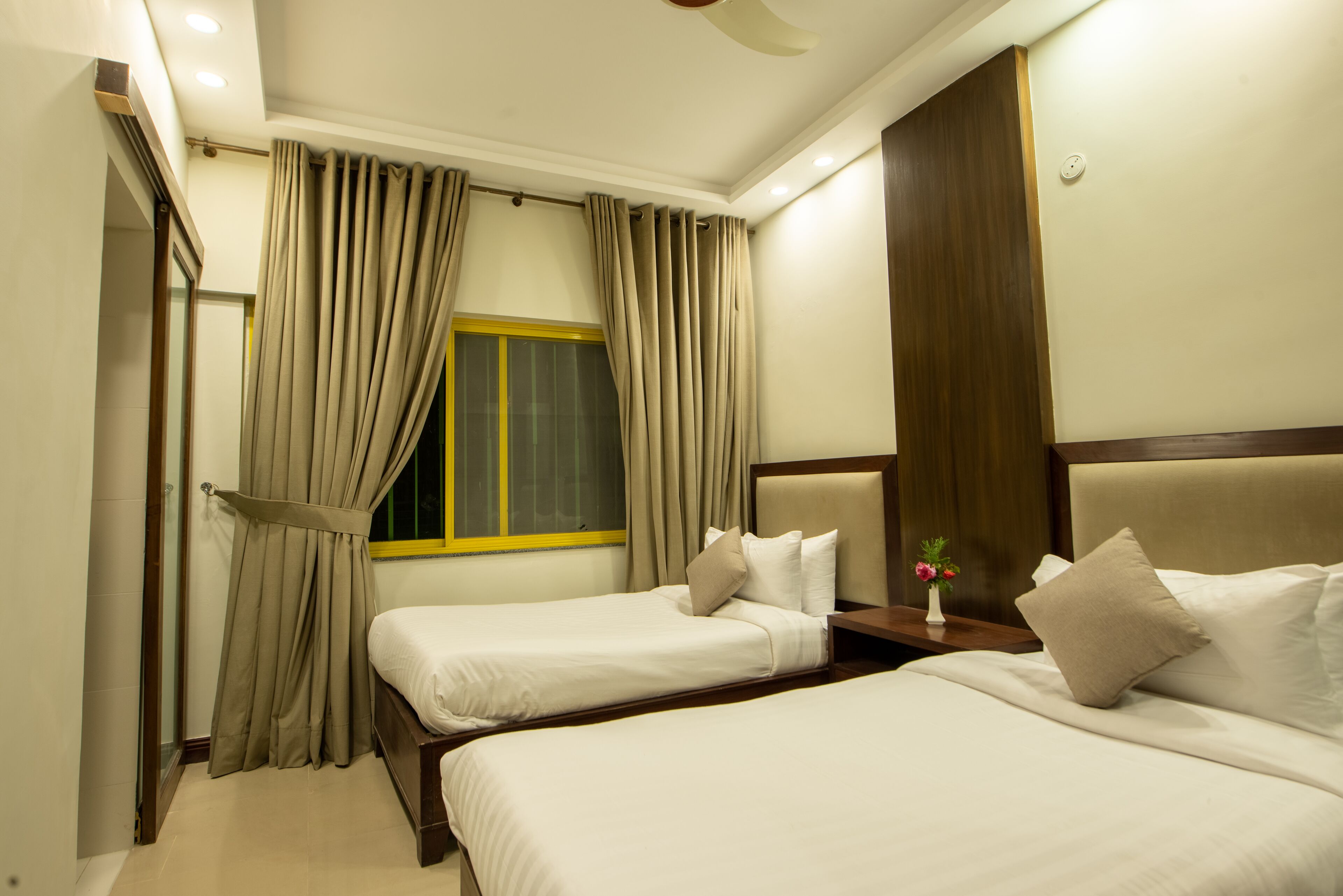 A hotel room with two beds, beige curtains, a window with yellow trim, a bedside table with a vase of flowers, and a wooden headboard.