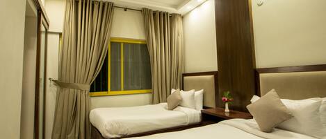 A hotel room with two beds, beige curtains, a window with yellow trim, a bedside table with a vase of flowers, and a wooden headboard.