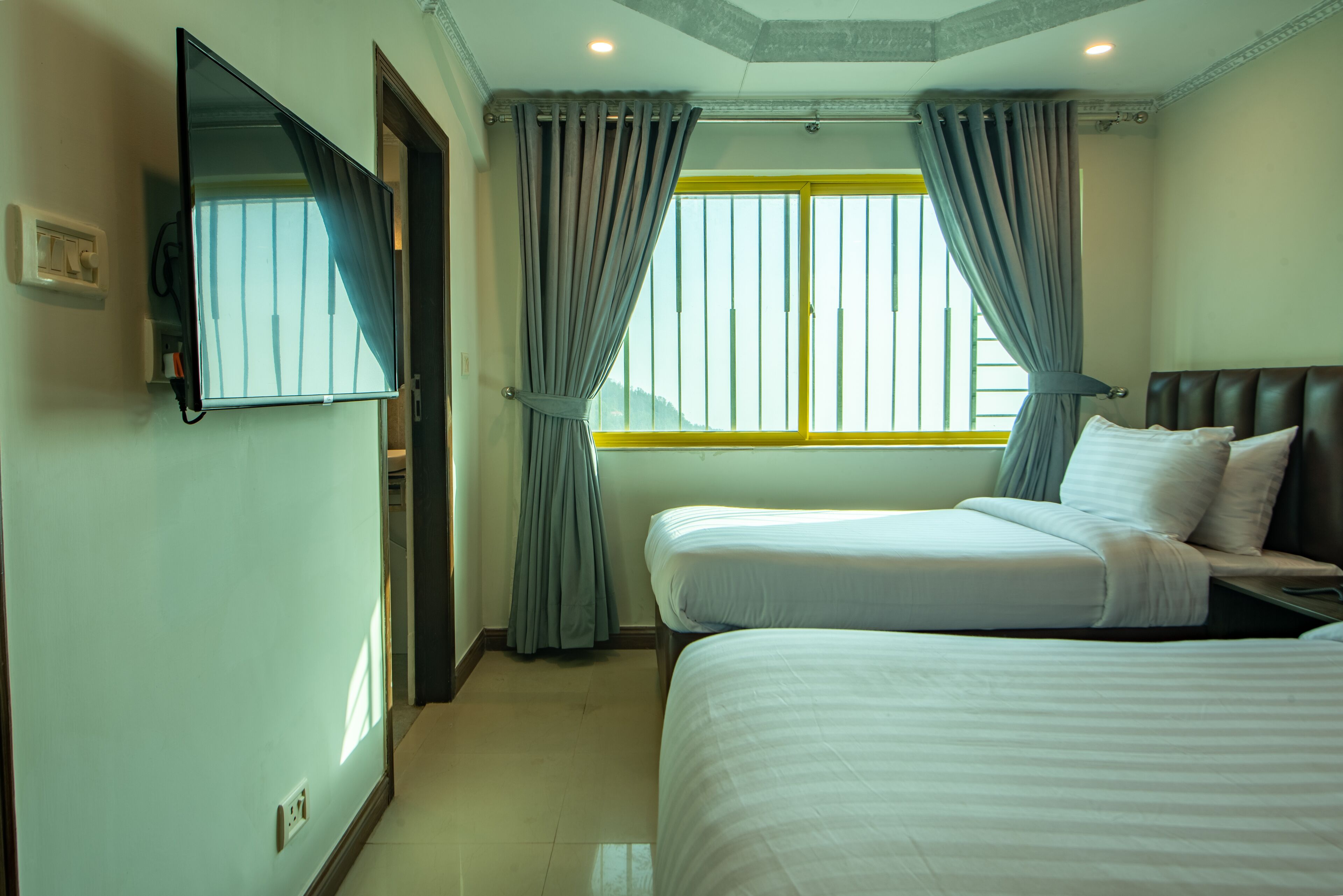 Habitación de hotel con dos camas, un televisor de pantalla plana montado en la pared, una ventana con cortinas y suelo de baldosas.