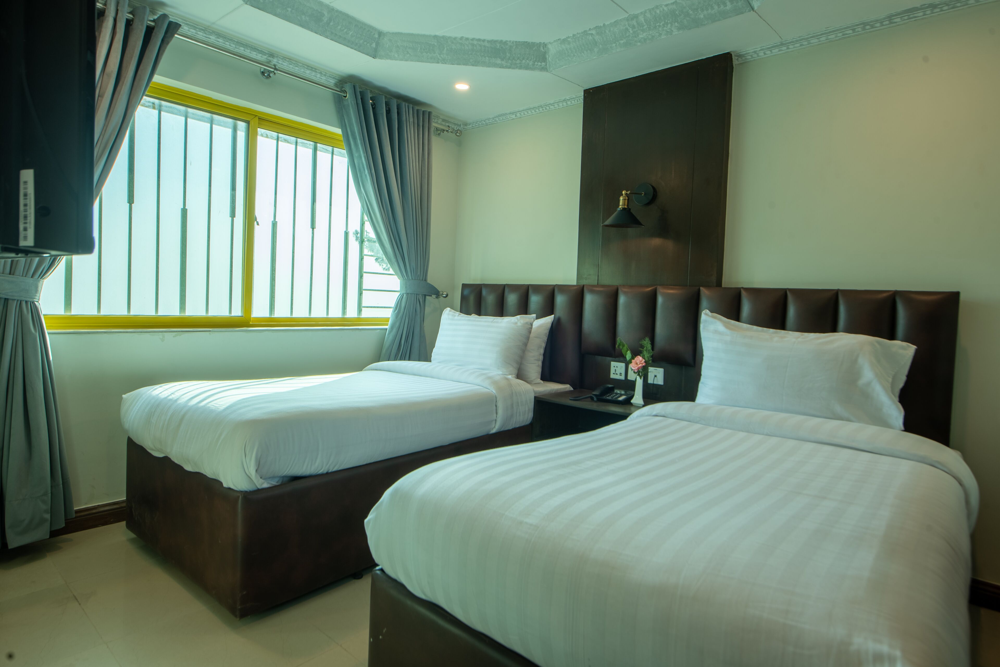 A hotel room with two beds, a large window with curtains, a bedside table with a lamp, and a wall-mounted light fixture.