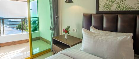 Habitación de hotel con cama, mesita de noche con un jarrón de flores, lámpara de pared y puerta corrediza de vidrio que da a un balcón con vista al mar.