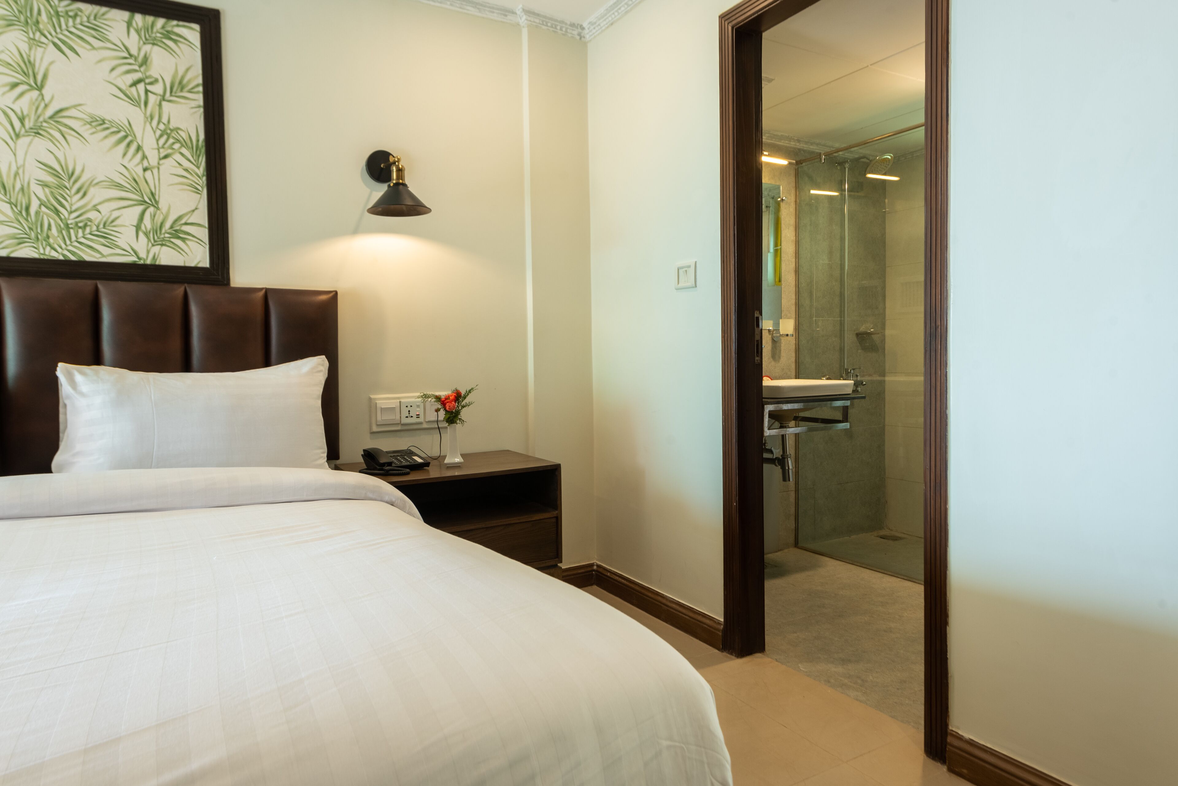 A hotel room with a large bed, a bedside table with a lamp, a nightstand with a phone and a vase of flowers, and a glass-enclosed shower area.