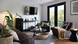 A modern living room with a sofa, armchair, and a coffee table. There is a TV mounted on the wall, a fireplace, and a large window. - Mi Casa Su Casa - stylish Interiors with Sauna! (Hepburn Springs)