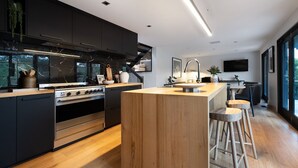 A modern kitchen with black cabinets, a wooden island, stainless steel appliances, and a large window providing a view of the outdoors. - Mi Casa Su Casa - stylish Interiors with Sauna! (Hepburn Springs)