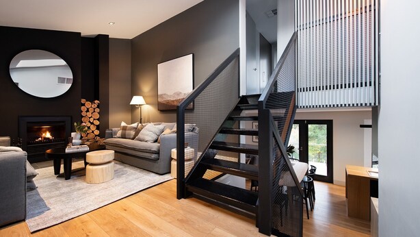 A modern living room with a fireplace, a grey sofa, a coffee table, and a staircase with a metal railing. - Mi Casa Su Casa - stylish Interiors with Sauna! (Hepburn Springs)