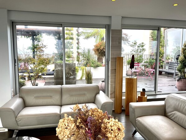 Un salon moderne avec un canapé beige, une porte vitrée donnant sur une terrasse extérieure et un vase rempli de fleurs séchées.